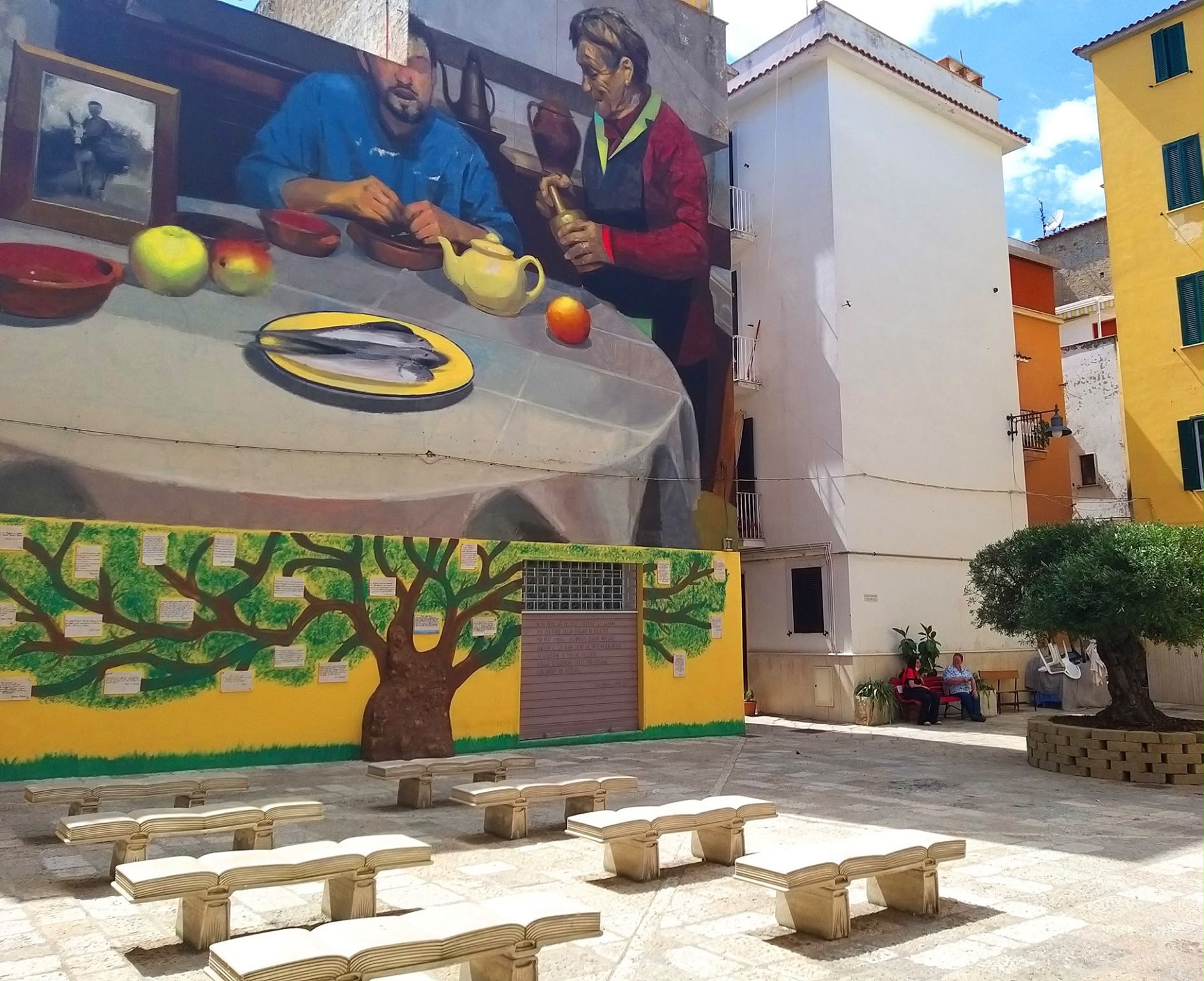 Piazzetta con un grande murale di una scena di cucina e panchine a forma di libro